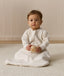 Infant wearing an ergoPouch Sleeping Bag 1.0 TOG Sleeved Oatmeal Marle, sitting on a white mat while holding a wooden toy.