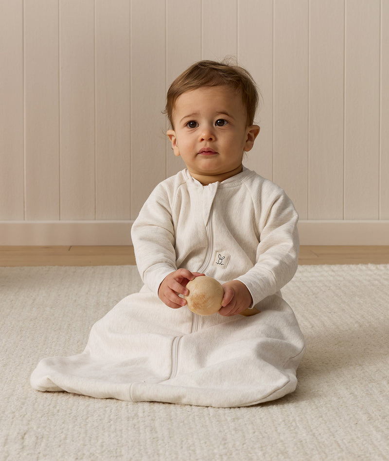 Infant wearing an ergoPouch Sleeping Bag 1.0 TOG Sleeved Oatmeal Marle, sitting on a white mat while holding a wooden toy.