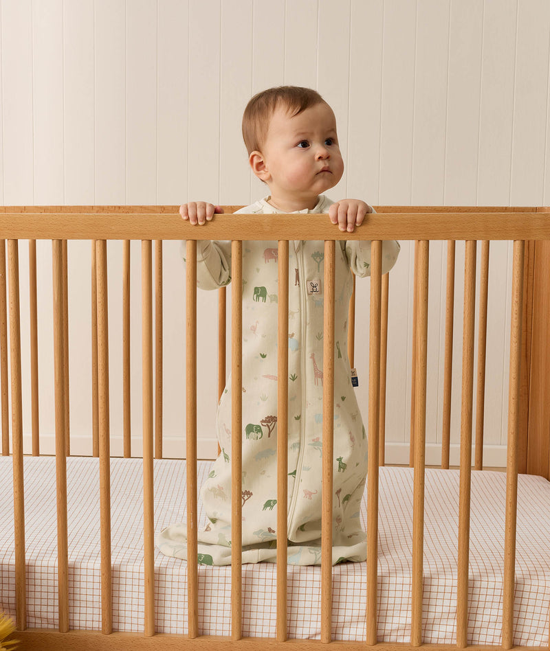 Infant standing inside a wooden cot wearing a long-sleeve 1.0 TOG Sleeping Bag with safari animal print