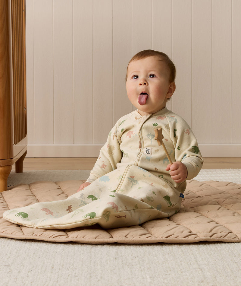 A playful baby sticking their tongue out while sitting on a beige quilted mat in a long-sleeved, animal-patterned sleeping bag and holding a wooden star toy.