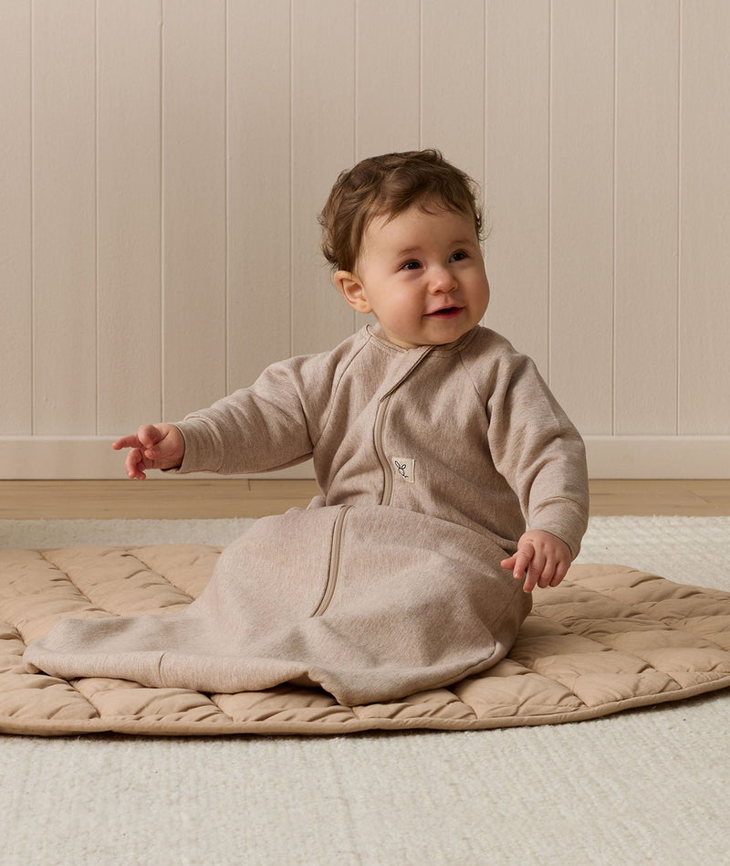 Infant wearing a long-sleeve Sleeping Bag 1.0 TOG Taupe Marle, sitting on a quilted mat