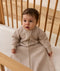 A baby wearing a long-sleeved beige Sleeping Bag 1.0 TOG, sitting upright inside a wooden cot with a grid-patterned sheet.