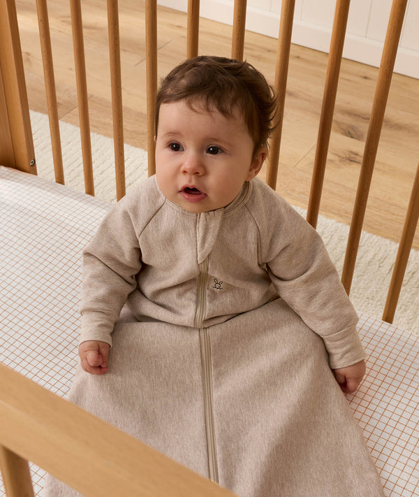 A baby wearing a long-sleeved beige Sleeping Bag 1.0 TOG, sitting upright inside a wooden cot with a grid-patterned sheet.