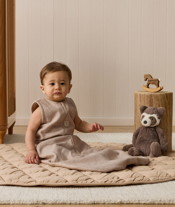 Infant wearing a sleeveless Sleeping Bag 1.0 TOG Taupe Marle, sitting on a quilted mat next to a stuffed animal.