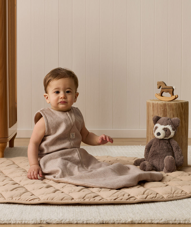 Infant wearing a sleeveless Sleeping Bag 1.0 TOG Taupe Marle, sitting on a quilted mat next to a stuffed animal.