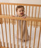 A smiling baby dressed in sleeveless Sleeping Bag, standing inside a wooden cot with a grid-patterned sheet.