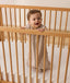 A smiling baby dressed in sleeveless Sleeping Bag, standing inside a wooden cot with a grid-patterned sheet.
