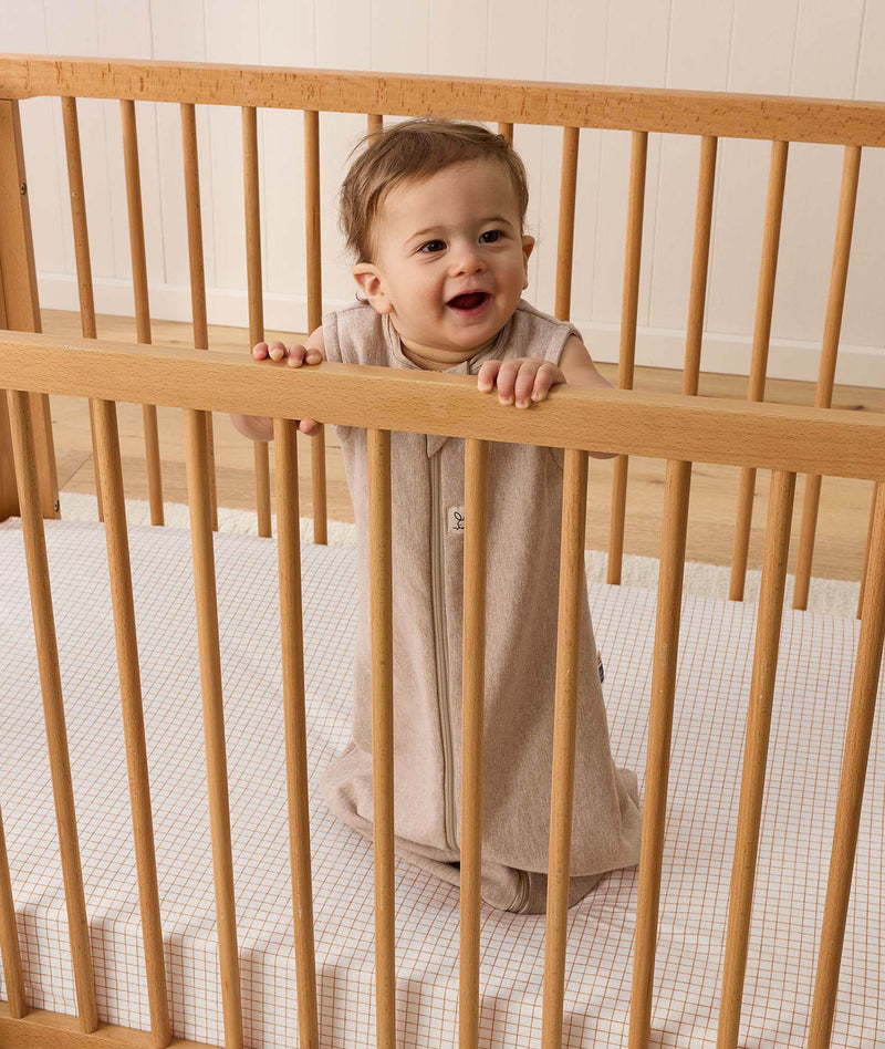 A smiling baby dressed in sleeveless Sleeping Bag, standing inside a wooden cot with a grid-patterned sheet.