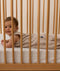 A smiling baby wearing a Taupe Marle Sleeping Bag, lying on their stomach inside a wooden cot with a grid-patterned sheet.