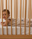 A smiling baby wearing a Taupe Marle Sleeping Bag, lying on their stomach inside a wooden cot with a grid-patterned sheet.