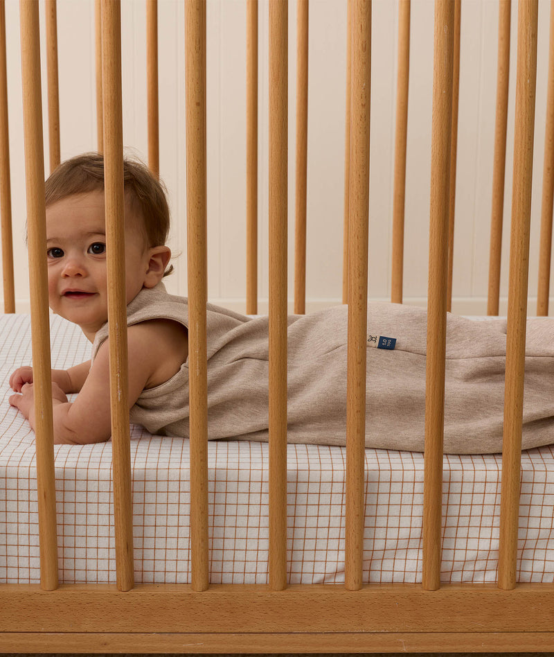 A smiling baby wearing a Taupe Marle Sleeping Bag, lying on their stomach inside a wooden cot with a grid-patterned sheet.