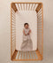 A happy baby lying in a wooden cot wearing a sleeveless Sleeping Bag 1.0 TOG Taupe Marle