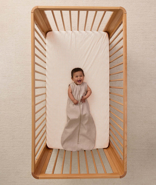 A happy baby lying in a wooden cot wearing a sleeveless Sleeping Bag 1.0 TOG Taupe Marle