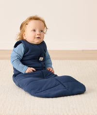 Baby sits on a rug, dressed in a dark blue ergoPouch 2.5 TOG Sleeping Bag and a Long Sleeve Layer underneath. 
