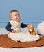 Infant wearing a cream Sleeping Bag 2.5 TOG with a blue long sleeve layer underneath, sitting next to a plush toy on a quilted mat.