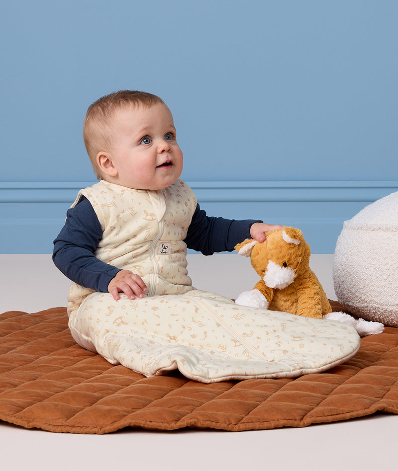 Infant wearing a cream Sleeping Bag 2.5 TOG with a blue long sleeve layer underneath, sitting next to a plush toy on a quilted mat.