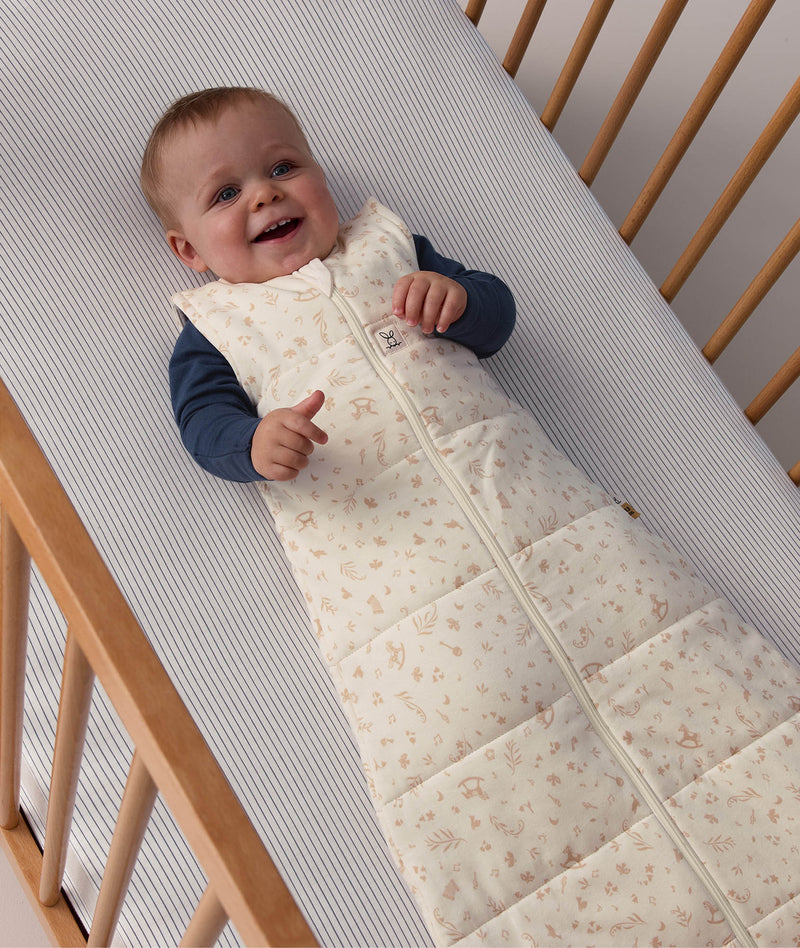 Smiling baby lying in a cot, wearing a patterned Sleeping Bag 2.5 TOG with a blue long sleeve layer underneath.
