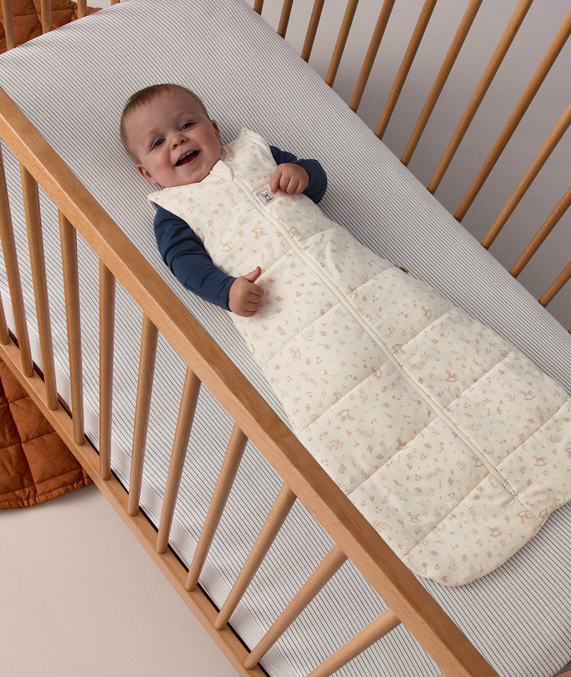 Smiling baby lying in a cot, wearing a patterned Sleeping Bag 2.5 TOG with a blue long sleeve layer underneath.