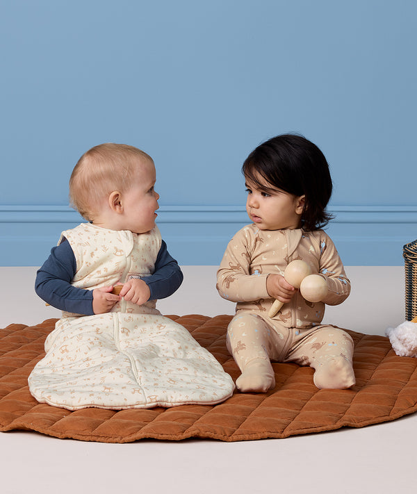 Two infants sitting on a play mat holding wooden toys, one wearing a cream-patterned Sleeping Bag 2.5 TOG and the other in a tan printed Long Sleeve Layer.