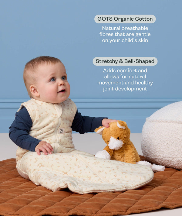 Infant wearing a cream Sleeping Bag 2.5 TOG, sitting next to a plush toy on a quilted mat with text highlighting organic cotton and stretchy bell-shaped design.