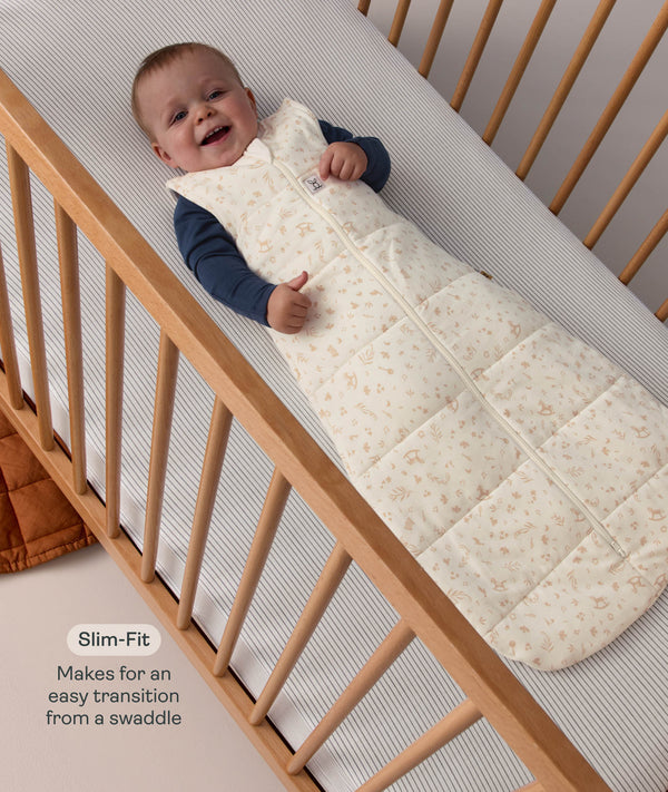 Smiling baby lying in a cot wearing a patterned ergoPouch Sleeping Bag 2.5 TOG with text highlighting slim-fit design for an easy transition from swaddling.