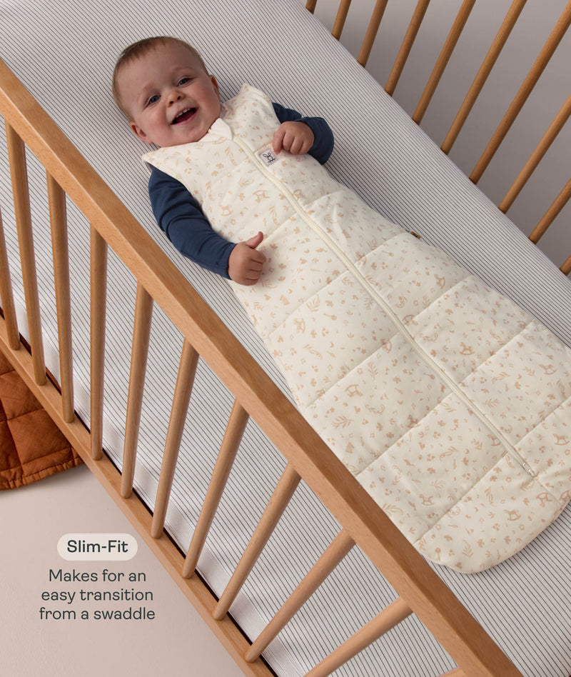 Smiling baby lying in a cot wearing a patterned ergoPouch Sleeping Bag 2.5 TOG with text highlighting slim-fit design for an easy transition from swaddling.
