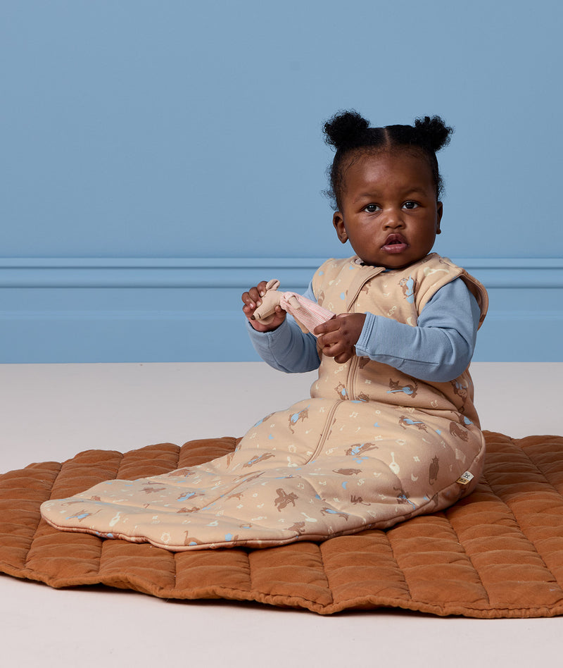Infant sitting on a brown mat, wearing a sleeveless Sleeping Bag 2.5 TOG with a long sleeve layer underneath.