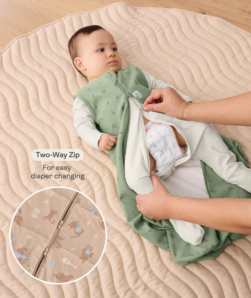 Baby lying down while adult unzips sage green Sleeping Bag using two-way zipper for nappy access, with a inset detail showing zipper close-up.