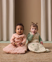 Two babies sits on a rug, each wearing ergoPouch Sleeping Bag in Elephant Parade and Savannah colourways.