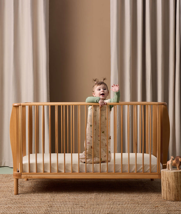 Baby standing in a crib wearing a 2.5 TOG Sleeping Bag with a subtle nature-themed pattern, including small green leaves and animals, over a green long-sleeve layer.