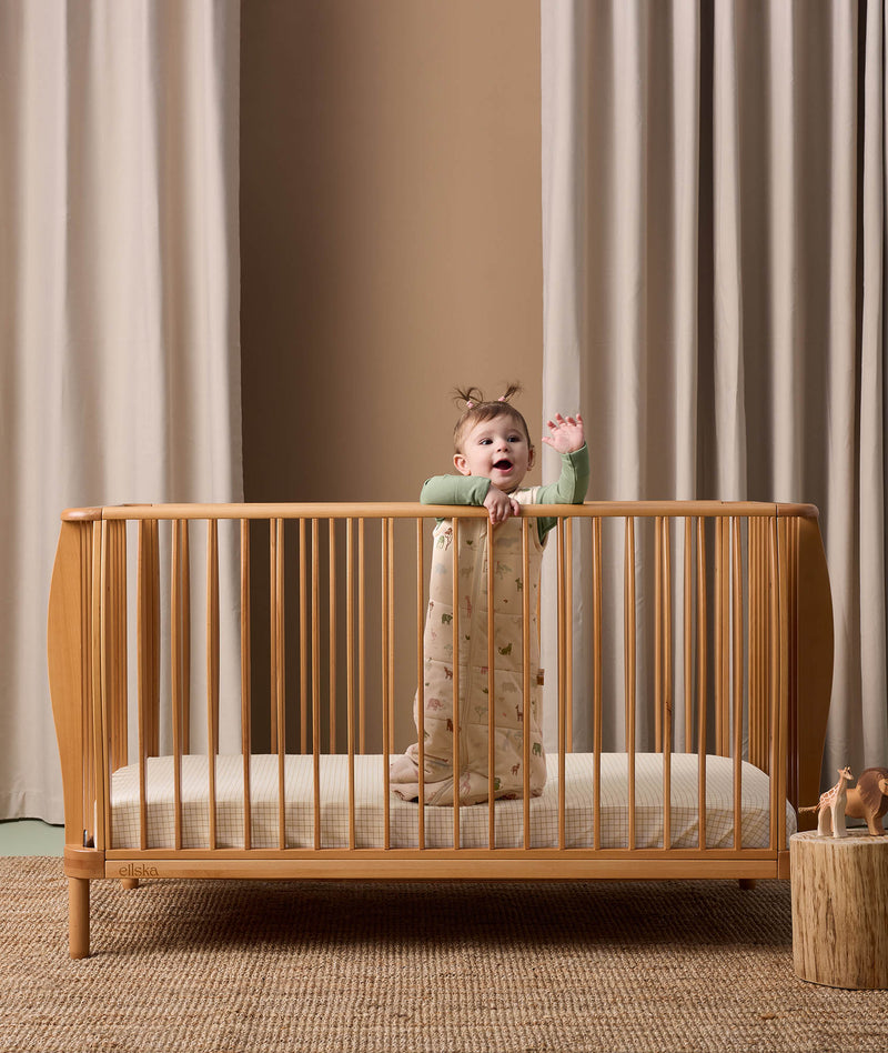 Baby standing in a crib wearing a 2.5 TOG Sleeping Bag with a subtle nature-themed pattern, including small green leaves and animals, over a green long-sleeve layer.