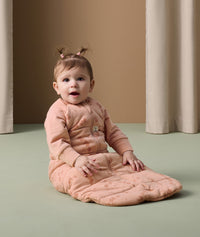An infant sits on the floor wearing an ergoPouch Sleeping Bag 3.5 TOG in blush pink colour with cute elephant prints.