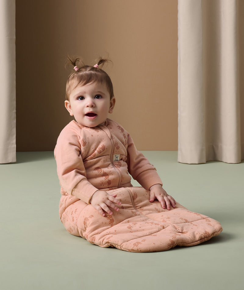 An infant sits on the floor wearing an ergoPouch Sleeping Bag 3.5 TOG in blush pink colour with cute elephant prints.