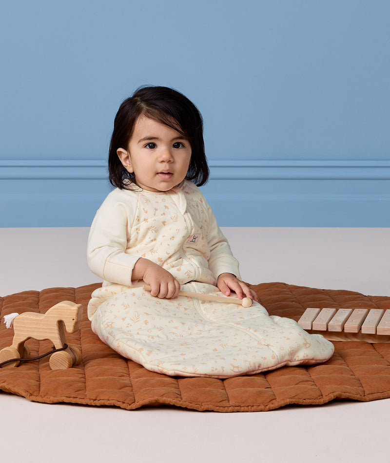 Infant wearing a sleeved Sleeping Bag 3.5 TOG, sitting on a quilted mat with wooden toys on the side