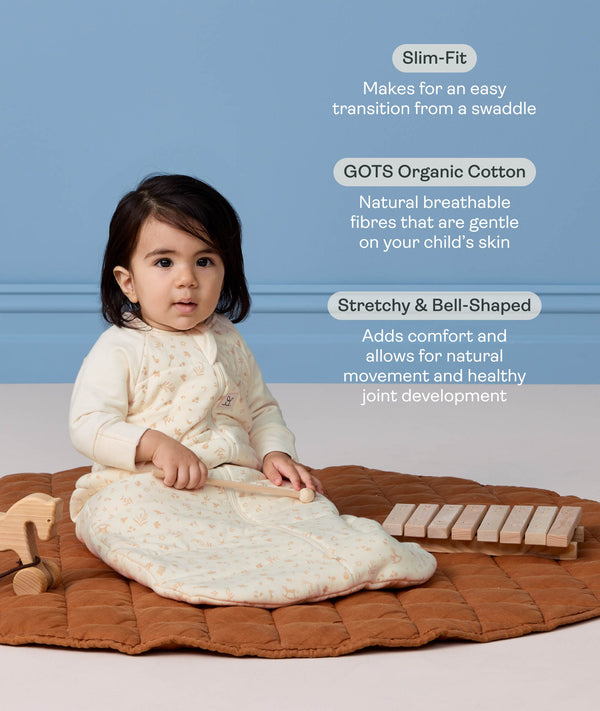 Infant sitting on a mat playing with a toy while wearing a cream patterned long-sleeve Sleeping Bag, with text highlighting its slim-fit, GOTS organic cotton, and stretchy bell-shaped design.