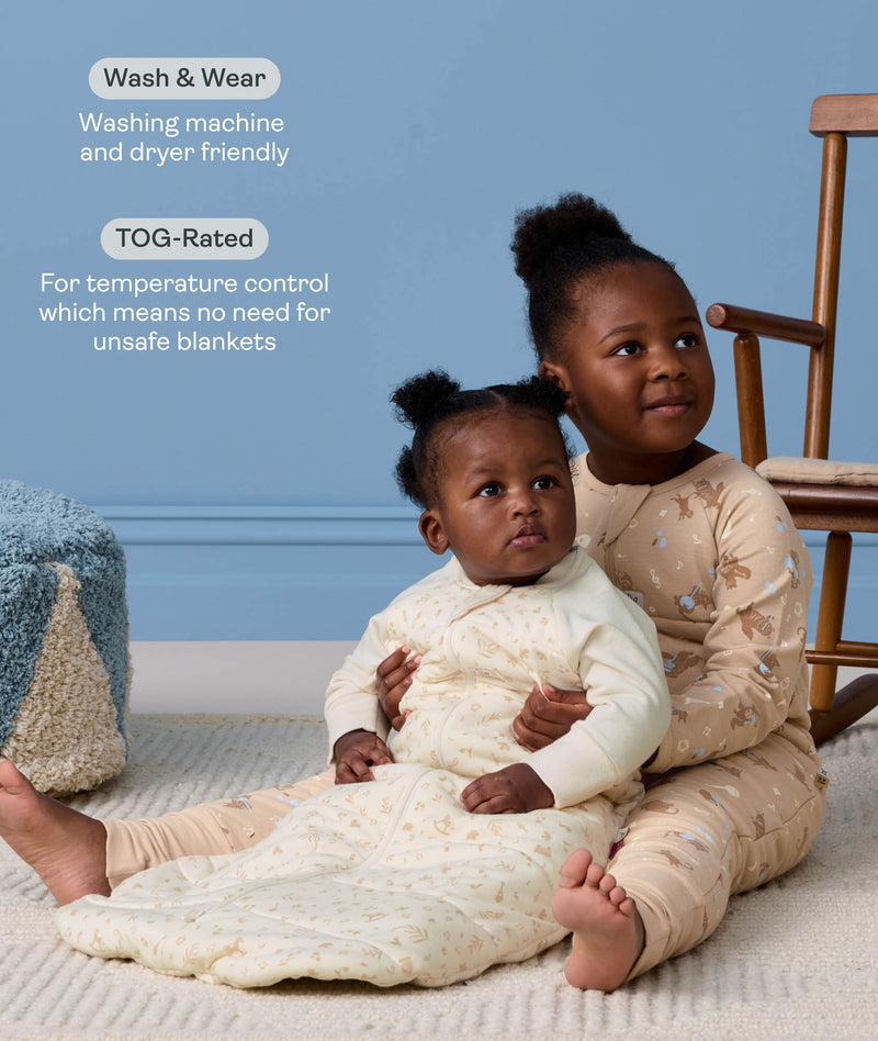 Preschooler sitting on a rug holding a baby wearing a cream patterned Sleeping Bag, with text highlighting wash-and-wear care and TOG ratings for temperature control.
