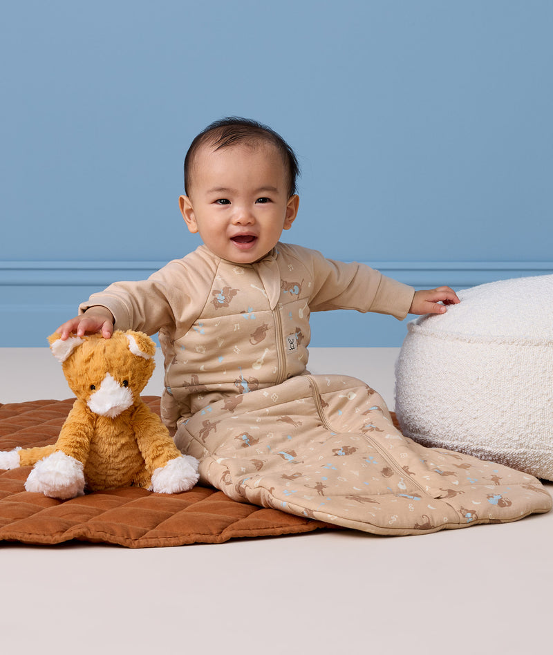 Infant wearing a sleeved Sleeping Bag 3.5 TOG, sitting next to a stuffed animal on a quilted mat.