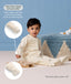 Toddler sitting on the floor playing a wooden xylophone in a cream patterned Sleep Onesie, with callouts demonstrating fold-over hand and foot cuffs for warmth and scratch protection.