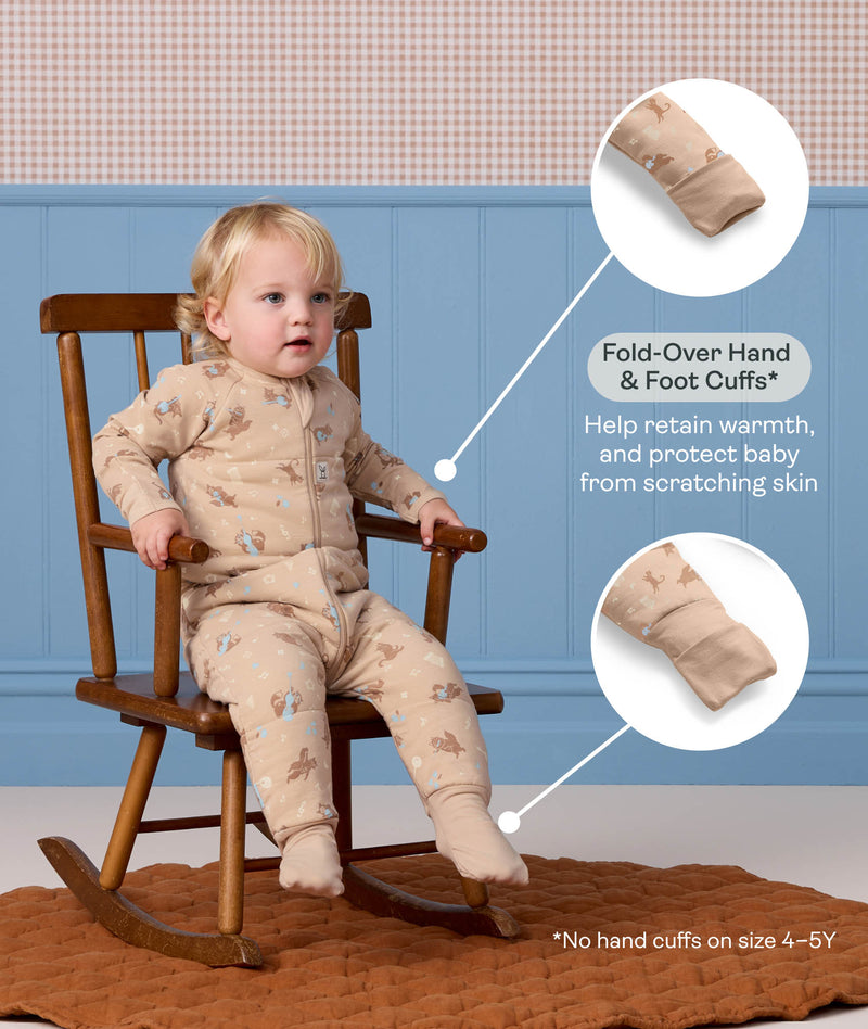 Toddler sitting in a wooden rocking chair wearing a beige patterned Sleep Onesie, with callouts highlighting fold-over hand and foot cuffs designed for warmth and scratch protection.