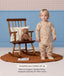 Toddler wearing a beige patterned Sleep Onesie standing next to a wooden rocking chair with a teddy bear, with text highlighting an Organic Toddler Pillow.