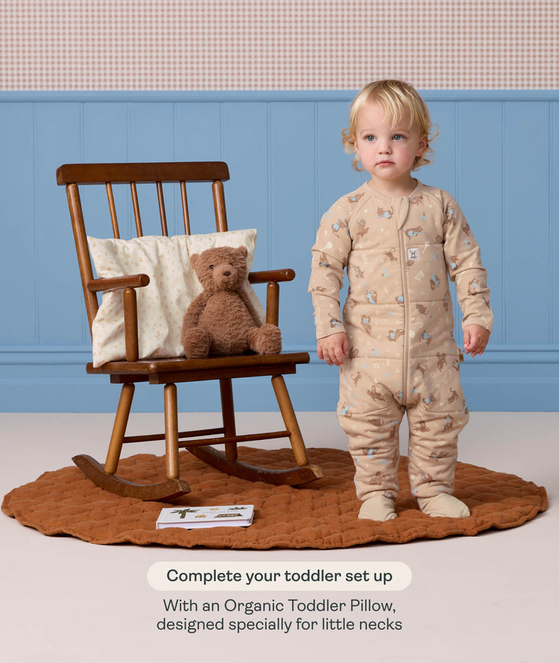 Toddler wearing a beige patterned Sleep Onesie standing next to a wooden rocking chair with a teddy bear, with text highlighting an Organic Toddler Pillow.