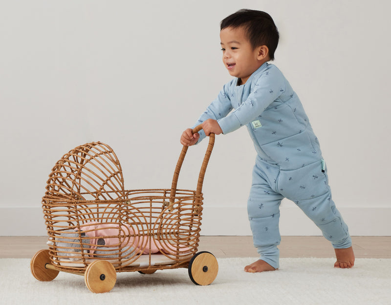 Toddler wearing a blue ergoPouch sleep onesie, pushing a wicker toy.
