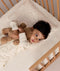 Toddler lying in a wooden cot on a cream patterned Organic Toddler Pillow while hugging a stuffed teddy bear.