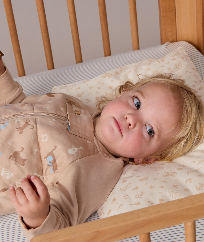 A toddler lies on their back in a wooden cot, resting their head on a patterned cream Organic Toddler Pillow while wearing a beige printed sleep suit.