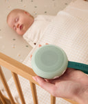 A parent holding a soft green portable white noise machine with a dark green strap labelled DRIFT AWAY, positioned next to a sleeping baby in a wooden crib.
