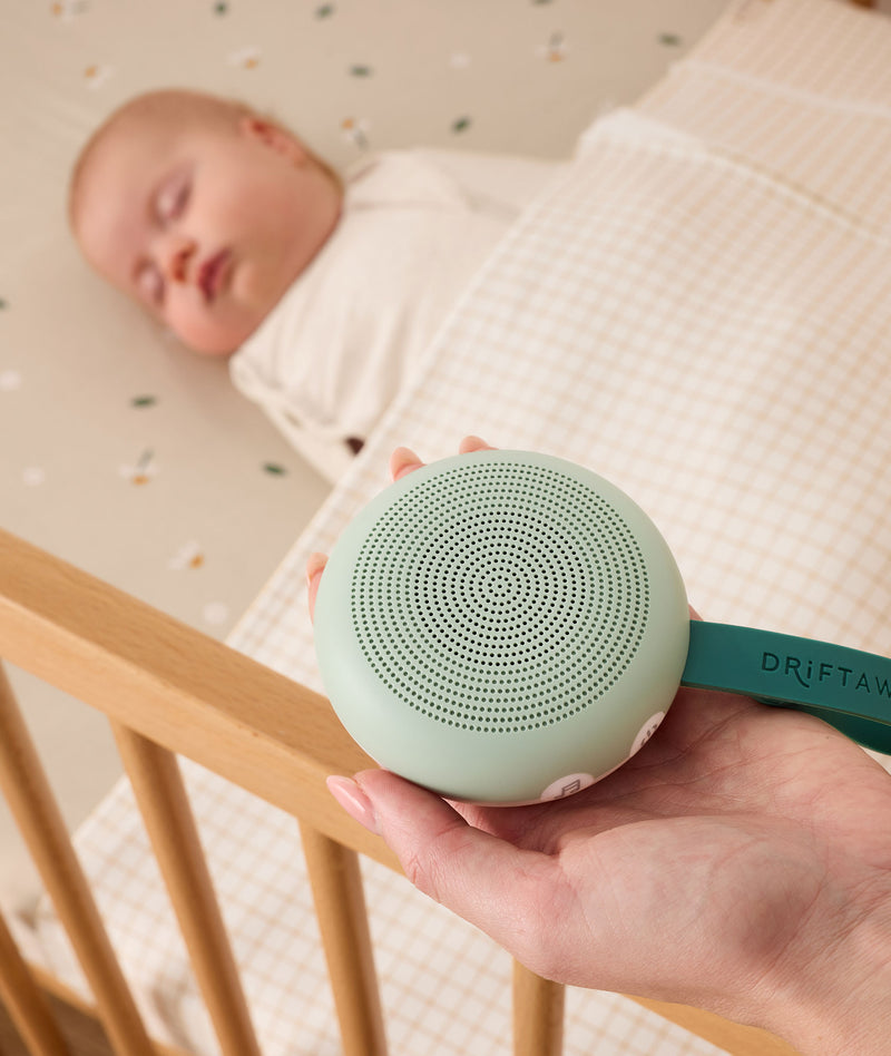 A parent holding a soft green portable white noise machine with a dark green strap labelled DRIFT AWAY, positioned next to a sleeping baby in a wooden crib.