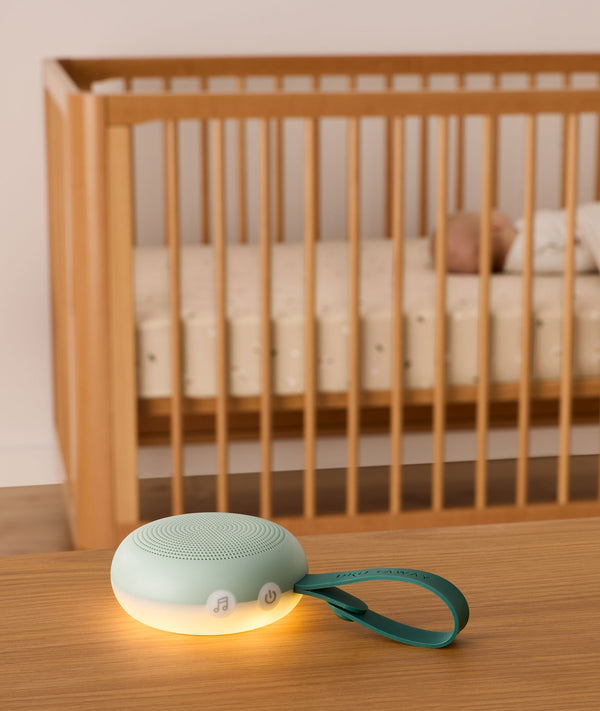 A soft green white noise machine with a glowing warm night light sits on a wooden table in the foreground. It features buttons, including music and power icons, and a green strap with the brand name “Drift Away.” In the background, a baby is peacefully sleeping in a wooden crib, emphasising the calming effect of the device.