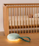 A sage green portable white noise machine with a glowing night light and dark green strap is placed on a wooden surface in the foreground, with a sleeping baby in a wooden crib visible in the background.