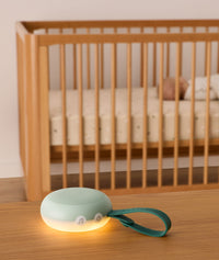 A sage green portable white noise machine with a glowing night light and dark green strap is placed on a wooden surface in the foreground, with a sleeping baby in a wooden crib visible in the background.