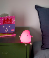 Pink glowing bedtime lamp shaped like a smiling character with small arms and legs, sitting on a green bedside table next to a children's birthday book.